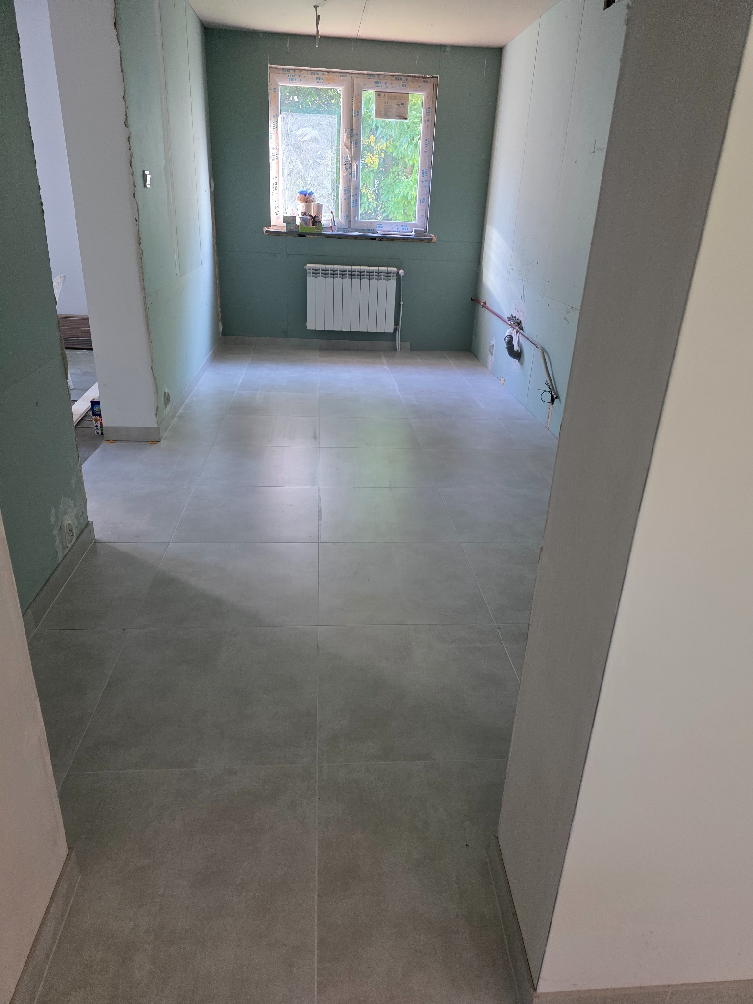 Wykończona podłoga z płytek w łazience w Krakowie, ściany przygotowane pod malowanie, widoczne instalacje. Grzejnik pod oknem.