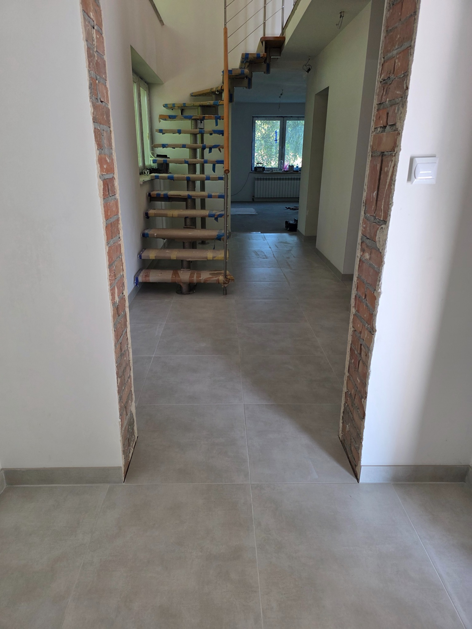Wnętrze w trakcie prac wykończeniowych: szare płytki podłogowe, fragment schodów zabezpieczonych folią, ściany z odsłoniętą cegłą przy przejściu.