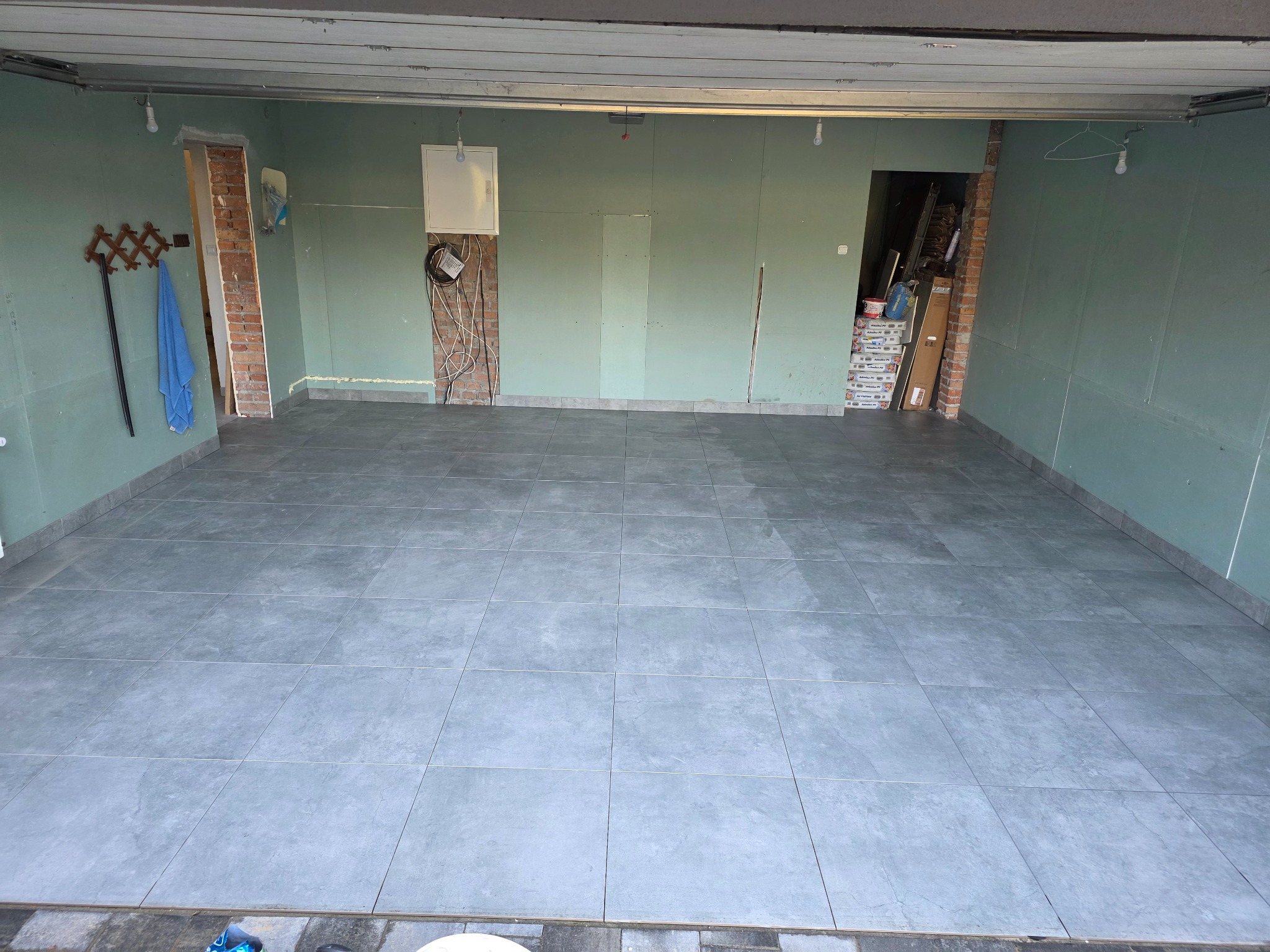 Wykończone podłogi z szarych płytek w pomieszczeniu z zielonymi ścianami z płyt kartonowo-gipsowych. Widoczne fragmenty instalacji elektrycznej i ceglanej obudowy drzwi.