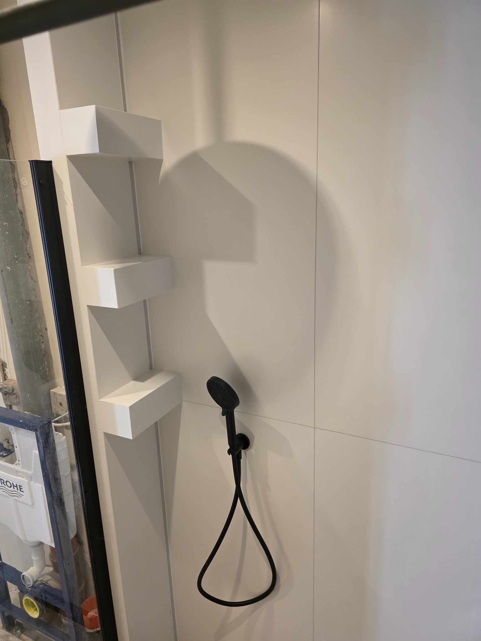 Minimalistyczna łazienka w trakcie wykańczania: białe panele ścienne, czarna słuchawka prysznicowa i półki w kształcie kostek. Widoczny stelaż podtynkowy.