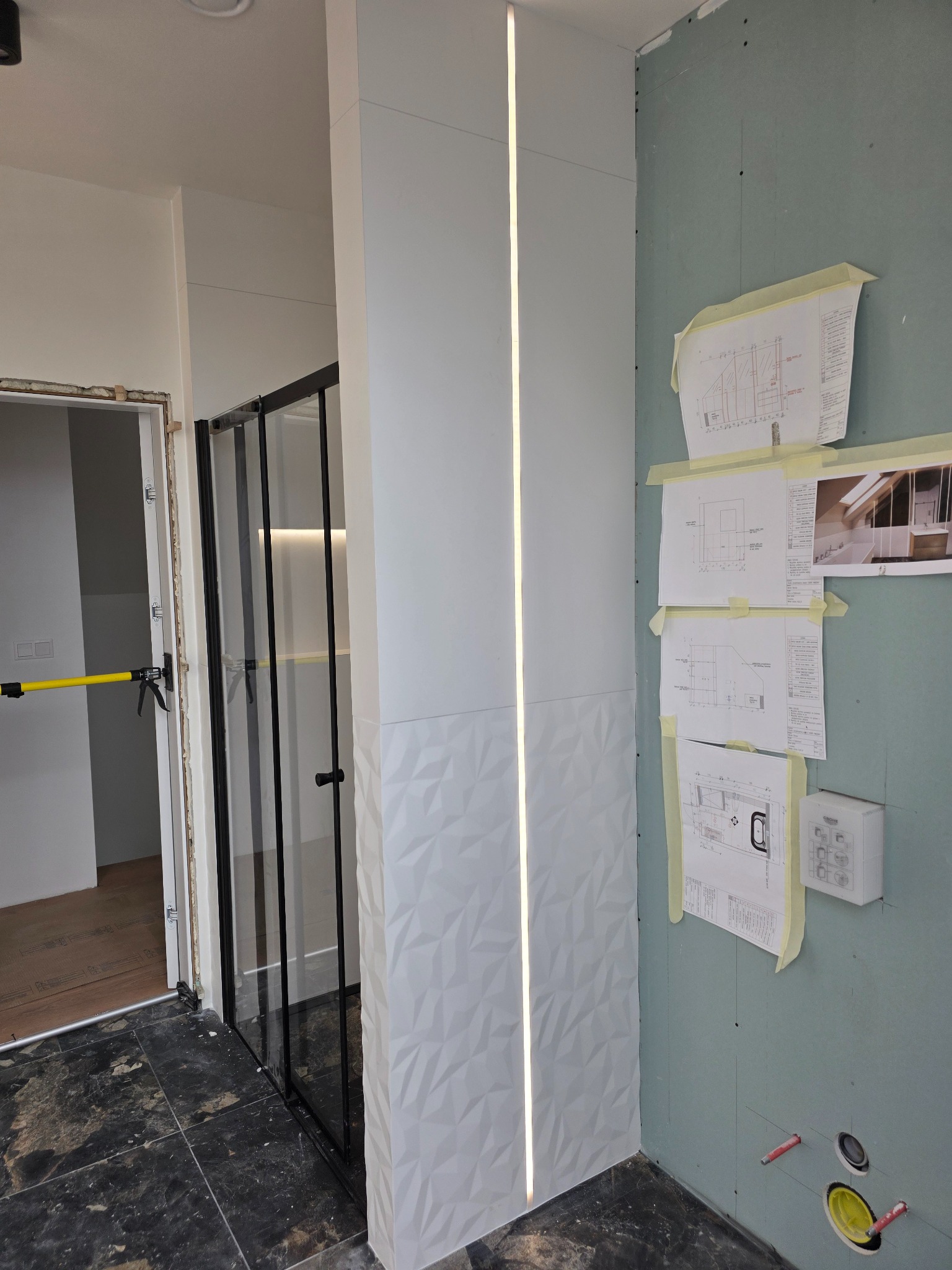 Łazienka w trakcie wykończenia: kabina prysznicowa z czarnymi ramami, ściana z płyt karton-gips i dekoracyjna z oświetleniem LED. Widoczne plany i instalacje.