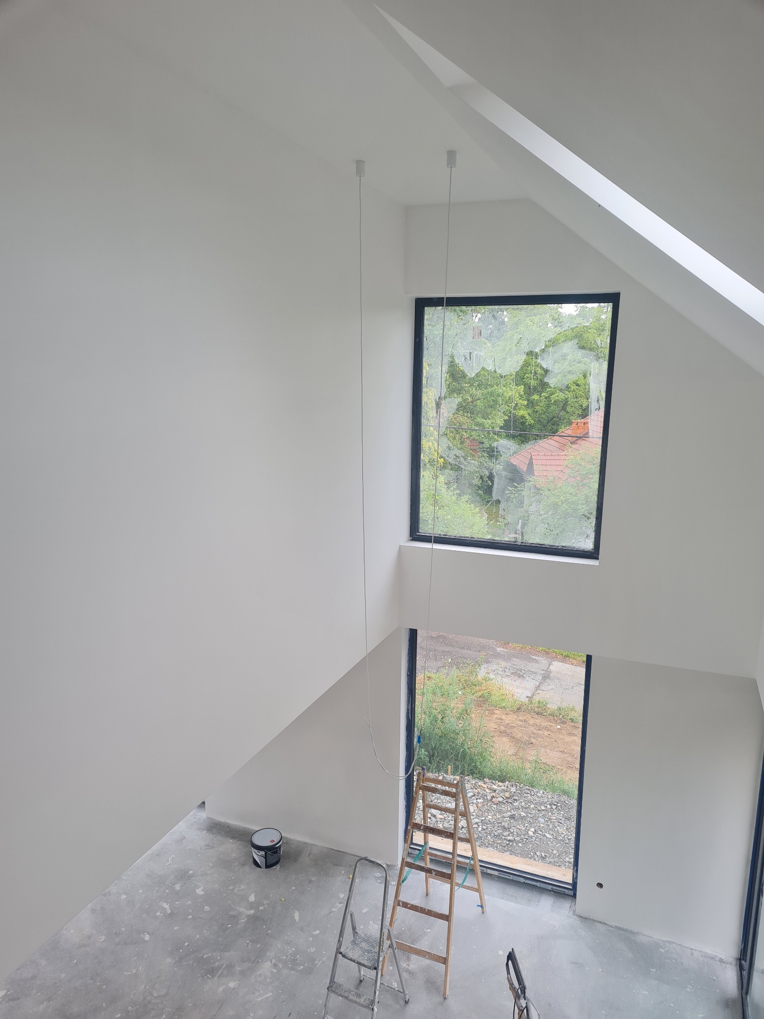 Wnętrze po malowaniu na biało, widoczne okna, drabiny malarskie i puszka farby. Podłoga zabezpieczona przed zabrudzeniem. Skosy sufitowe.