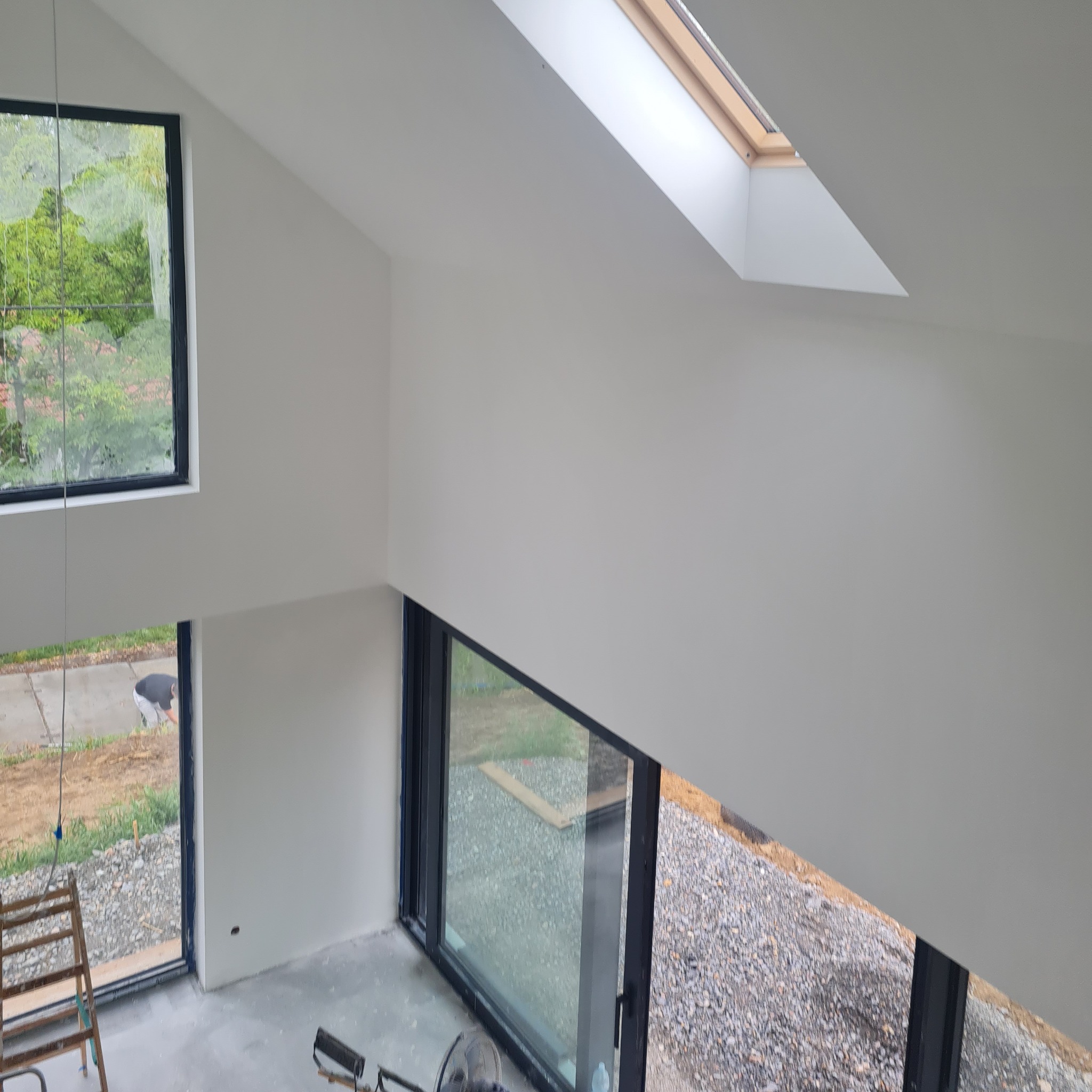 Wnętrze po malowaniu na biało, z oknem dachowym i dużymi oknami z czarnymi ramami. Widoczny fragment drabiny i podłoga przygotowana do dalszych prac.