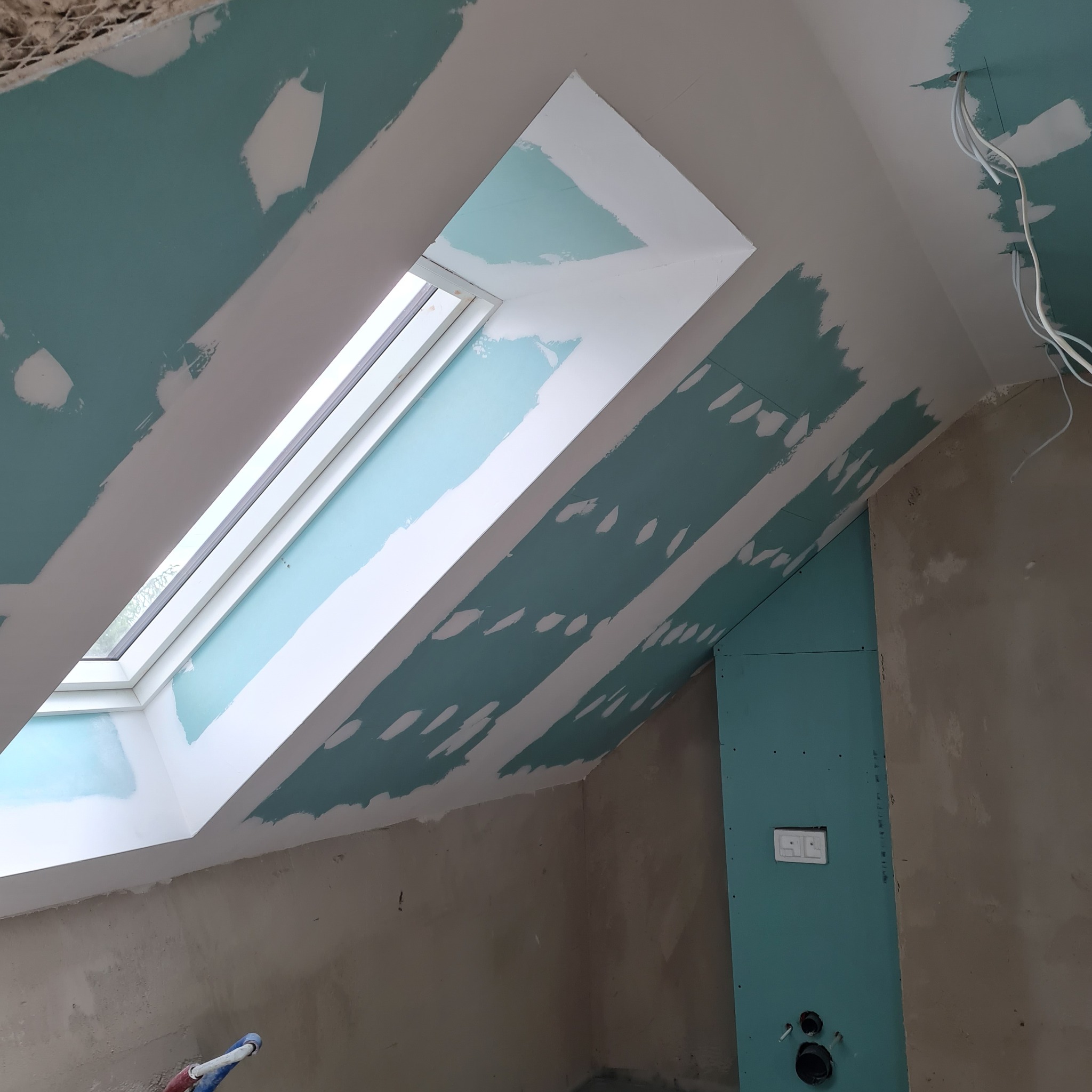 Poddasze z oknem dachowym w trakcie prac wykończeniowych: szpachlowanie łączeń płyt gipsowo-kartonowych na skosach i ścianach. Widoczne instalacje elektryczne.