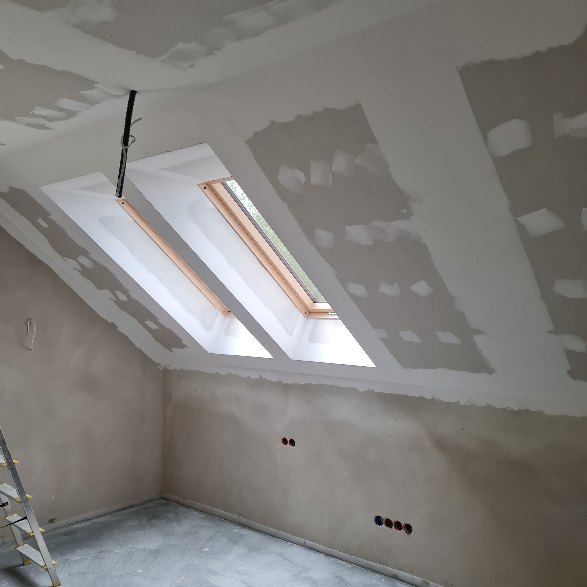 Pokój na poddaszu w trakcie prac wykończeniowych: ściany i sufit po szpachlowaniu, widoczne okna dachowe i instalacja elektryczna. Fragment drabiny w lewym dolnym rogu.