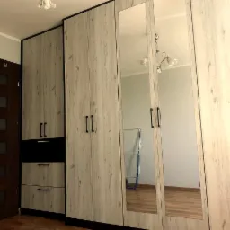 Zestaw szaf z frontami imitującymi drewno, z czarnymi akcentami i lustrami wbudowanymi w drzwi, odbijającymi wnętrze pokoju z suszarką na ubrania.