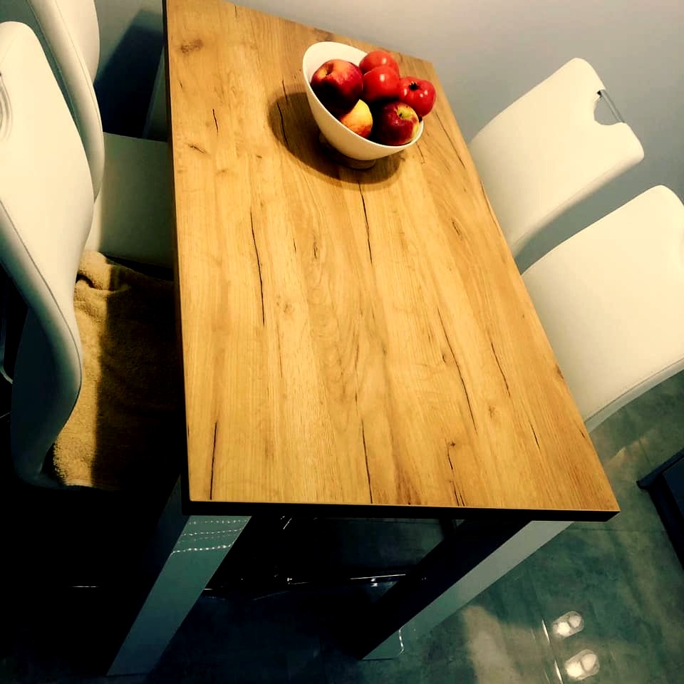 Stół z blatem z jasnego drewna o wyraźnym usłojeniu, na metalowych nogach, z misą czerwonych jabłek i białymi krzesłami w tle.
