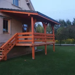 Nowo wybudowany drewniany taras z zadaszeniem i schodami, przylegający do domu z szarą elewacją, widok na trawnik i drzewa w tle.