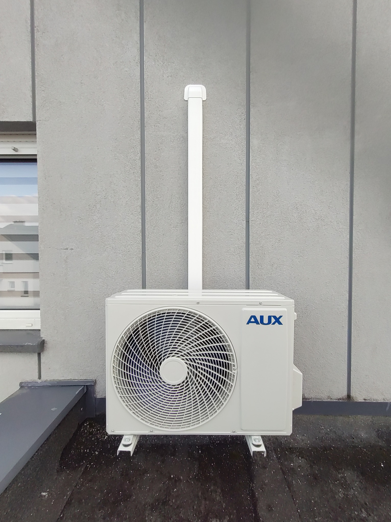 Zewnętrzna jednostka klimatyzacji AUX zamontowana na szarym budynku, widoczna rura odprowadzająca skropliny.