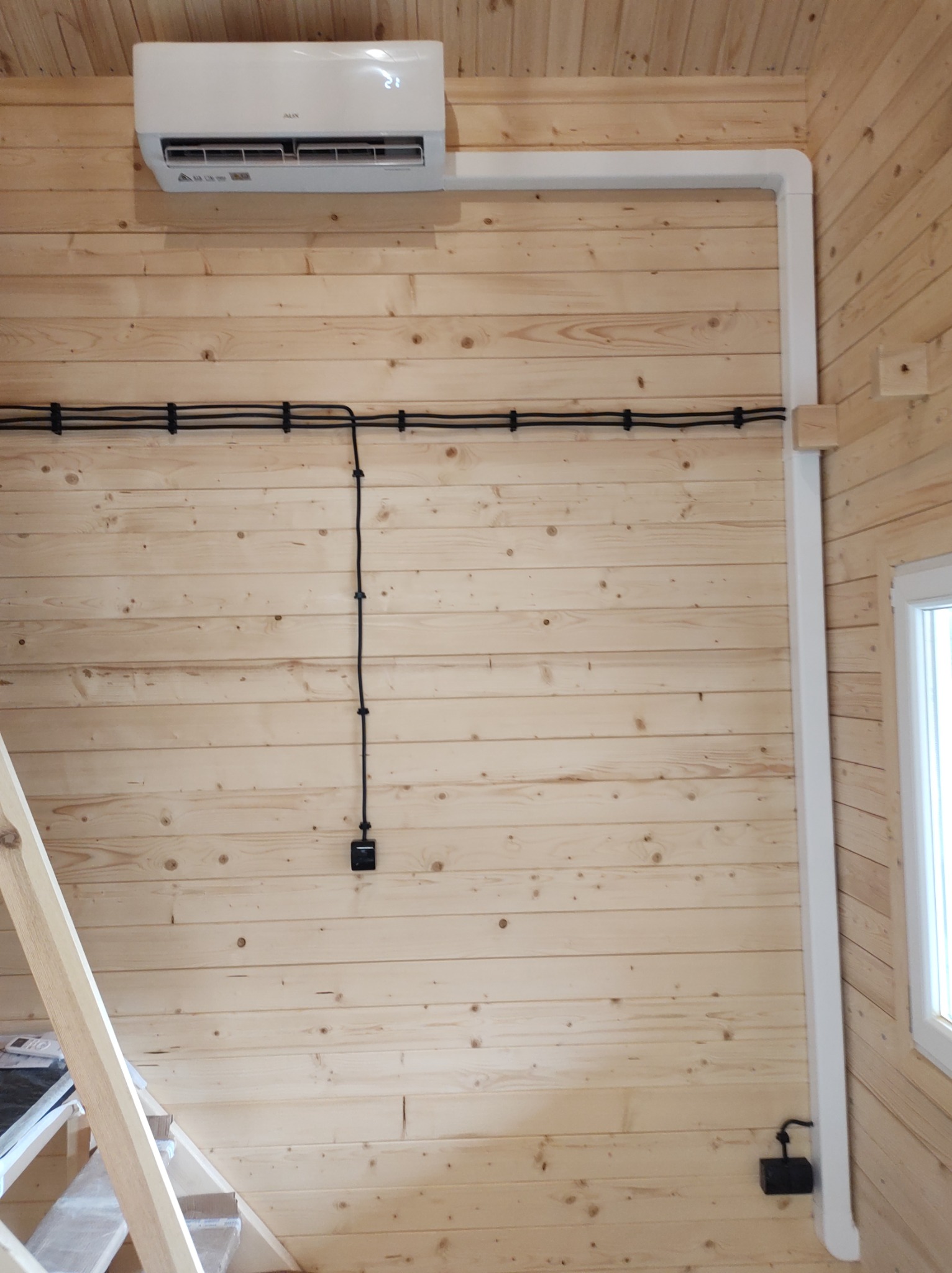 Wnętrze domu wykończonego drewnem z zamontowanym klimatyzatorem AUX, widoczne rury odprowadzające i instalacja elektryczna doprowadzona po ścianie.