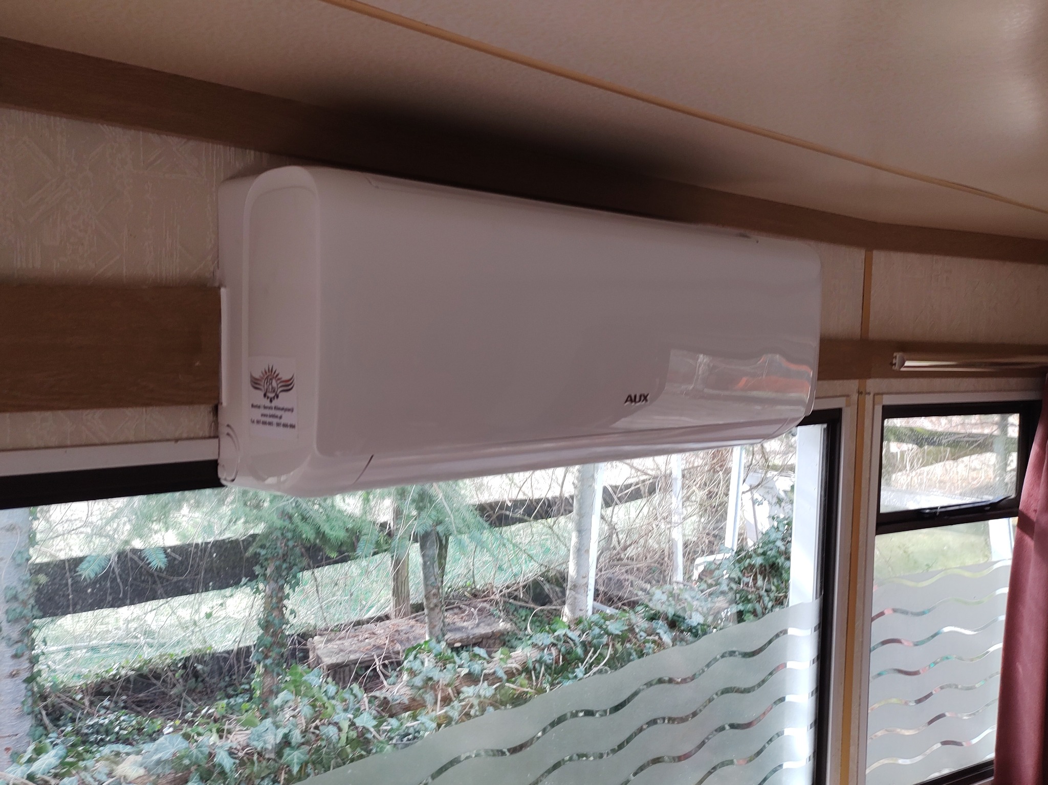 Biała jednostka wewnętrzna klimatyzacji marki AUX zamontowana nad oknem z folią dekoracyjną, widok na zarośnięty ogród.