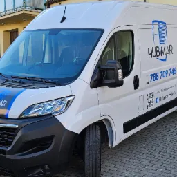 Biała furgonetka firmy Hubmar z niebieskim logo i numerem telefonu zaparkowana na brukowanej ulicy przed budynkiem, oferująca usługi montażu okien, drzwi, rolet i bram.
