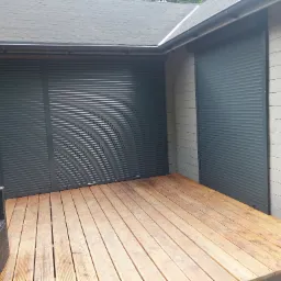 Szare rolety zewnętrzne zamknięte na oknach budynku z drewnianą elewacją i tarasem, widoczny fragment czarnego fotela.