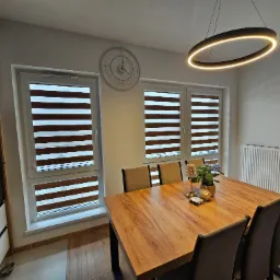 Jadalnia z drewnianym stołem, krzesłami i trzema oknami z roletami dzień i noc, okrągła lampa sufitowa i zegar na ścianie.