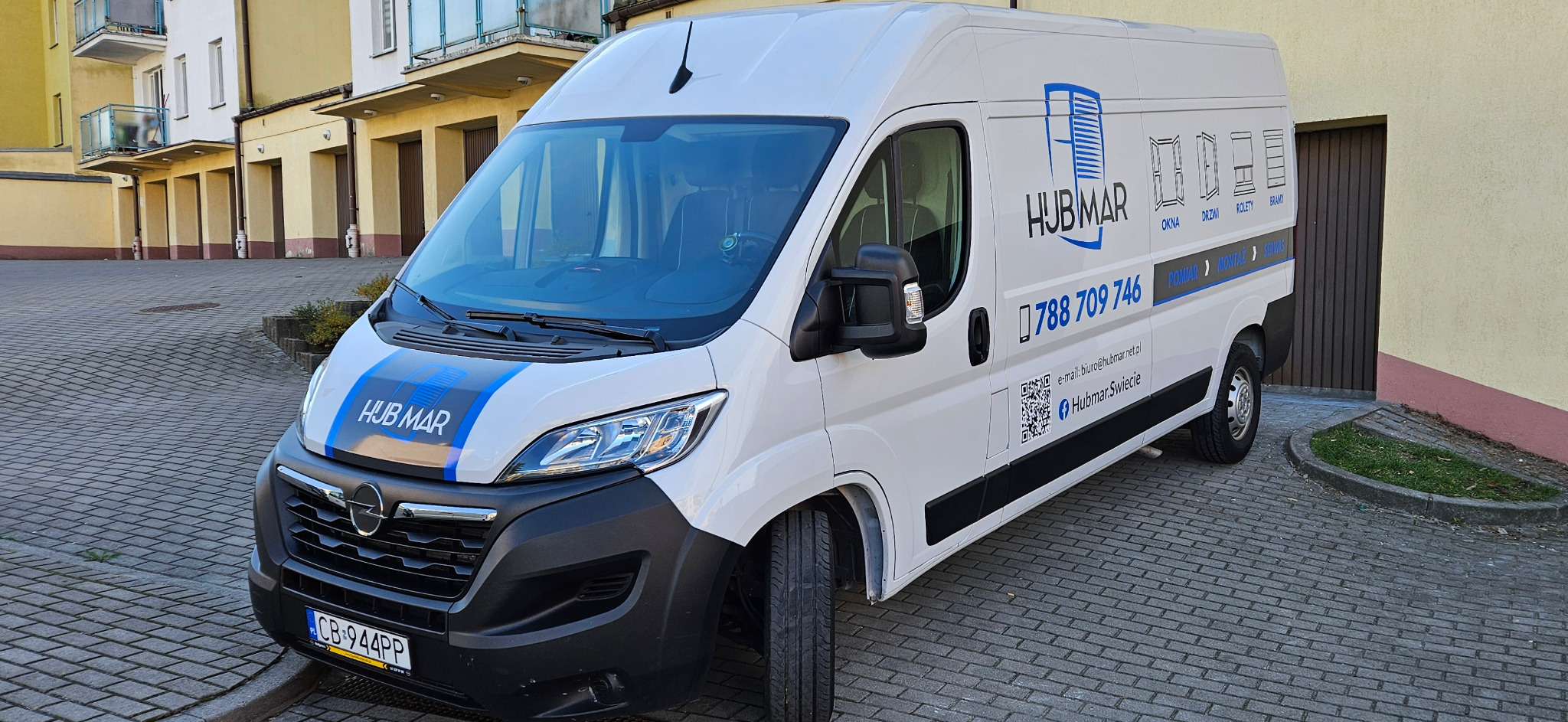Biała furgonetka firmy Hubmar z niebieskim logo i numerem telefonu zaparkowana na brukowanej ulicy przed budynkiem, oferująca usługi montażu okien, drzwi, rolet i bram.