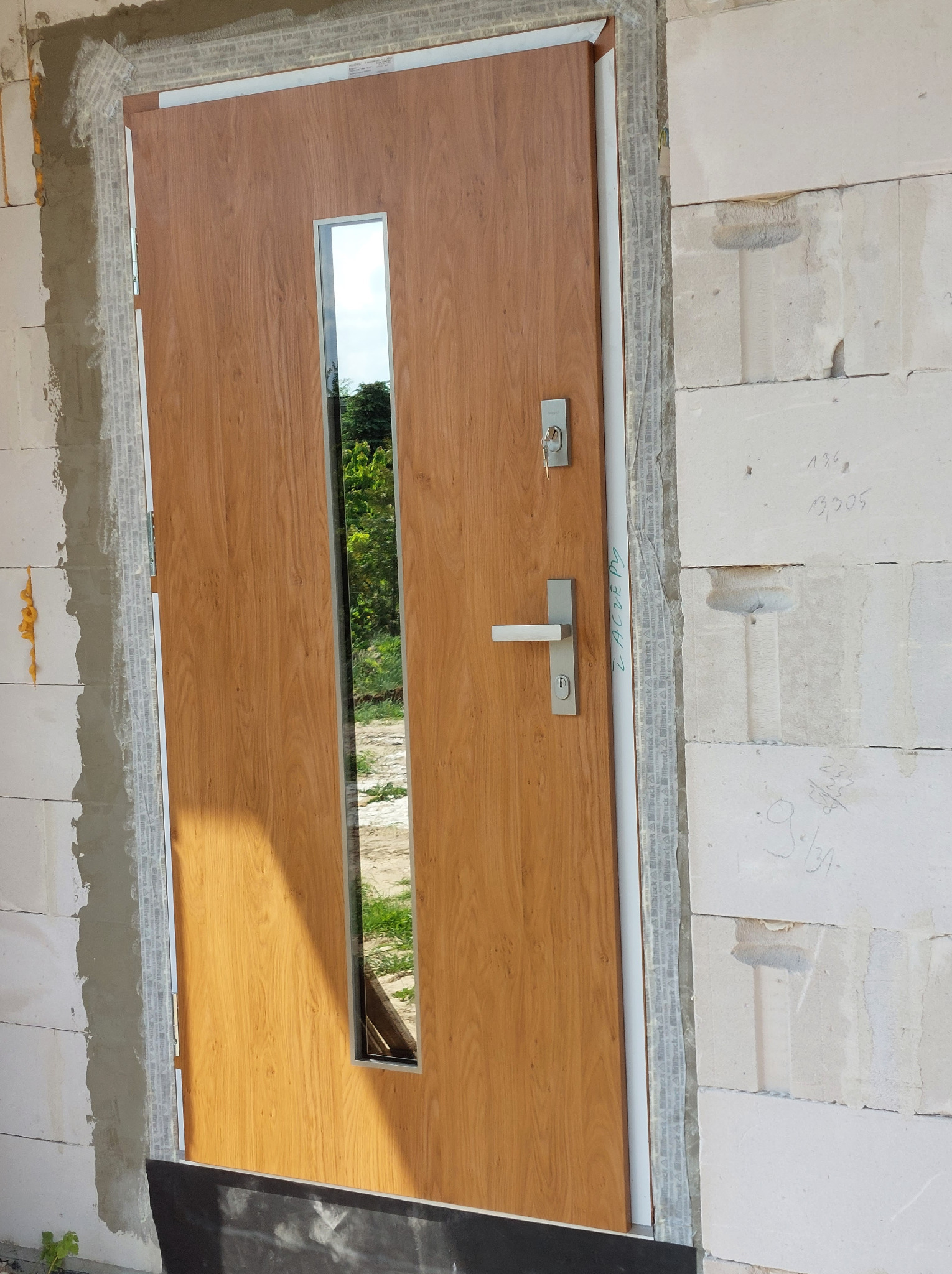 Montaż drzwi wejściowych z przeszkleniem, widoczne bloczki betonowe i pianka montażowa wokół ościeżnicy, klucz w zamku.