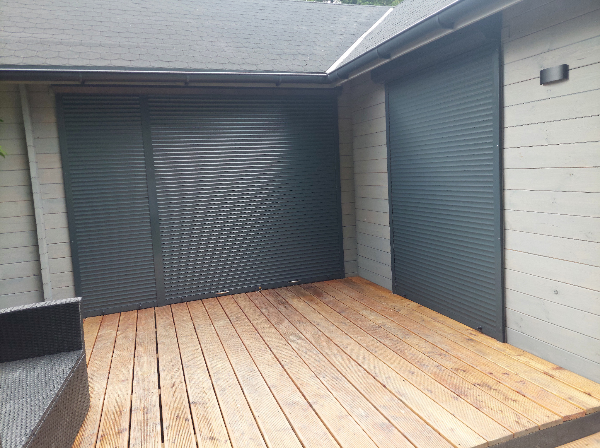 Szare rolety zewnętrzne zamknięte na oknach budynku z drewnianą elewacją i tarasem, widoczny fragment czarnego fotela.
