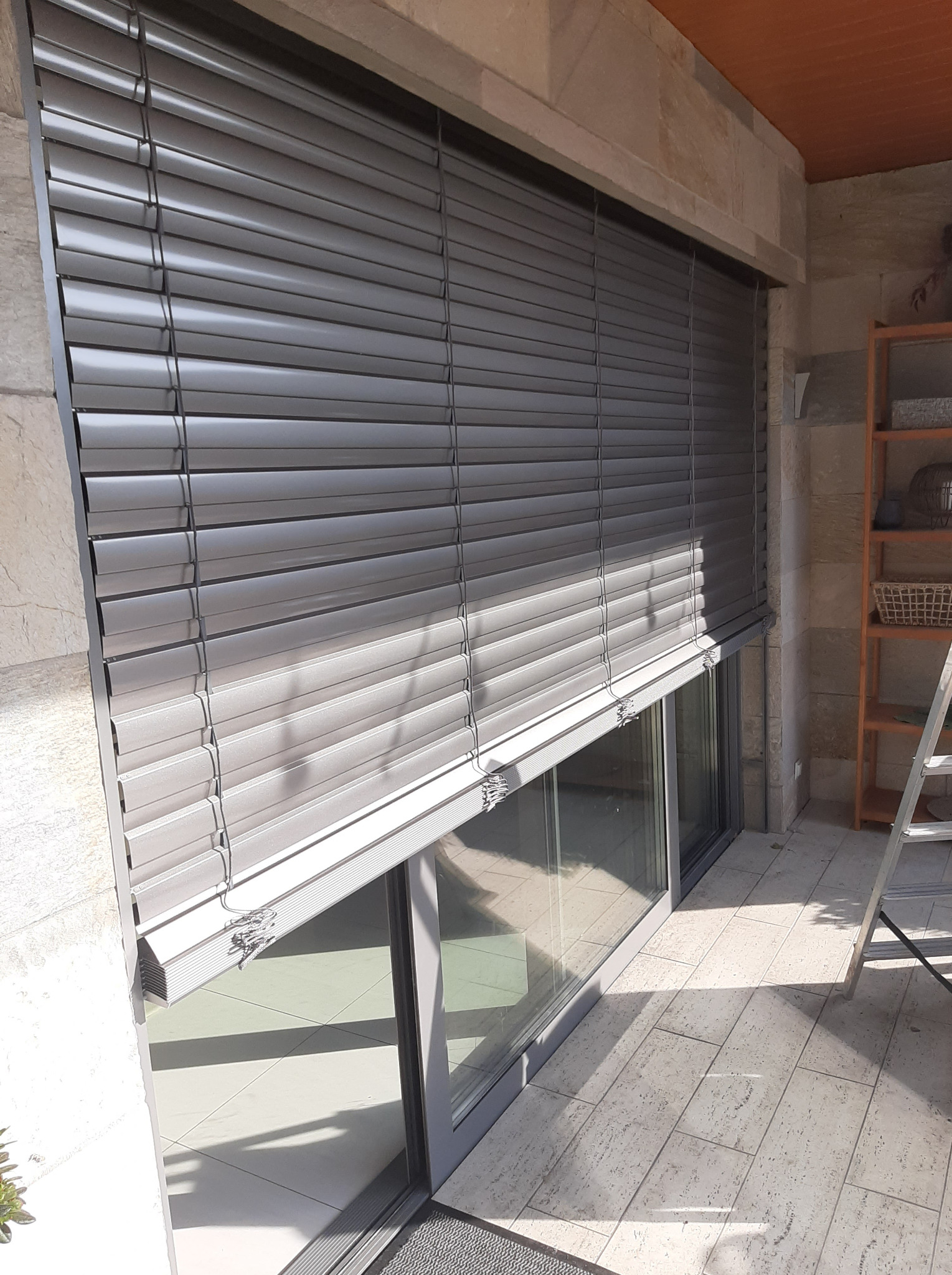 Zamknięte szare żaluzje fasadowe zamontowane na oknie balkonowym, widoczne z zewnątrz, z elementami dekoracyjnymi z kamienia i drabina w tle.