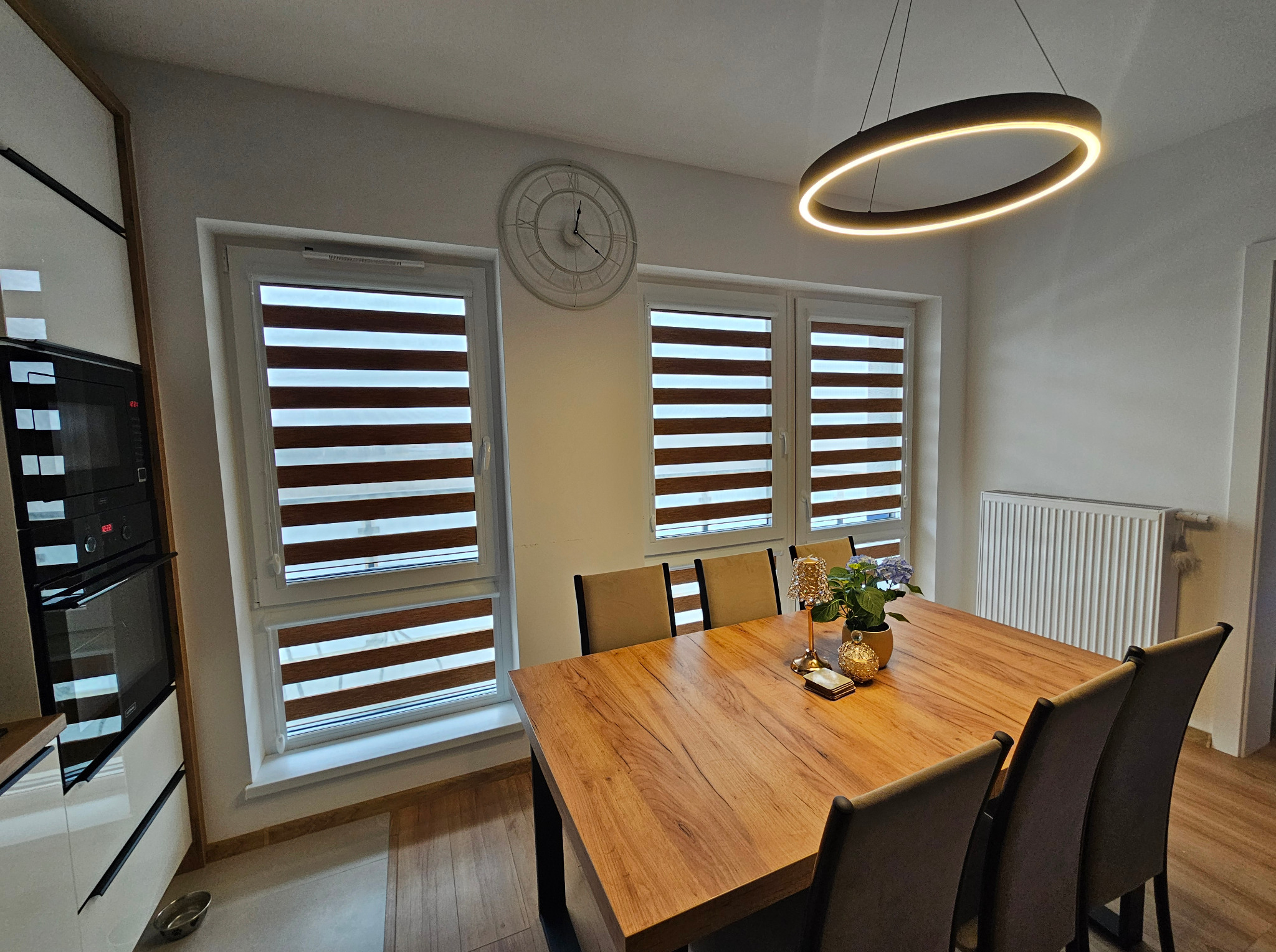 Jadalnia z drewnianym stołem, krzesłami i trzema oknami z roletami dzień i noc, okrągła lampa sufitowa i zegar na ścianie.