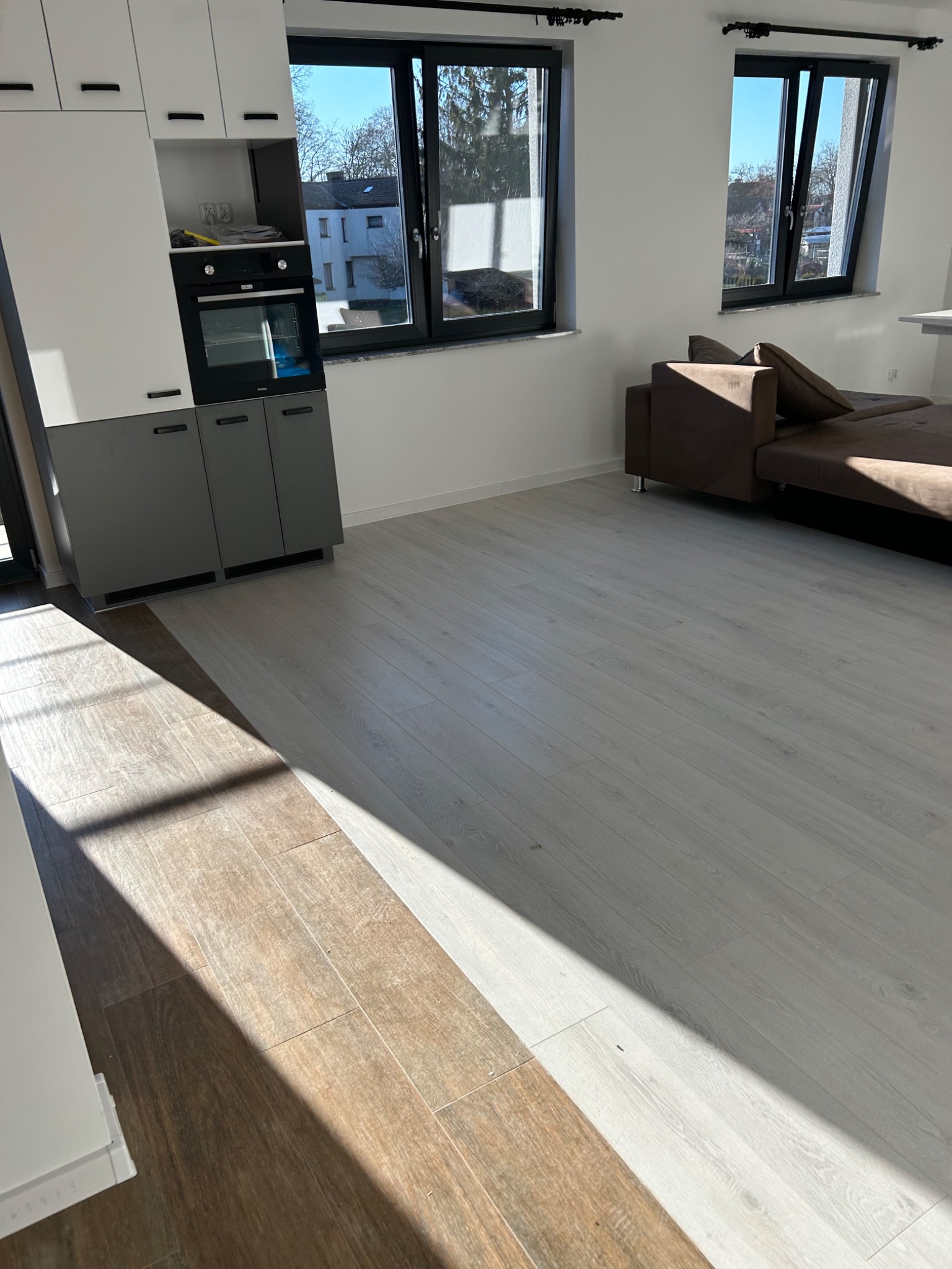 Jasne panele podłogowe w nowym mieszkaniu, z widocznym fragmentem brązowej sofy i zabudową kuchenną z piekarnikiem, oświetlone silnym światłem słonecznym wpadającym przez duże okna.