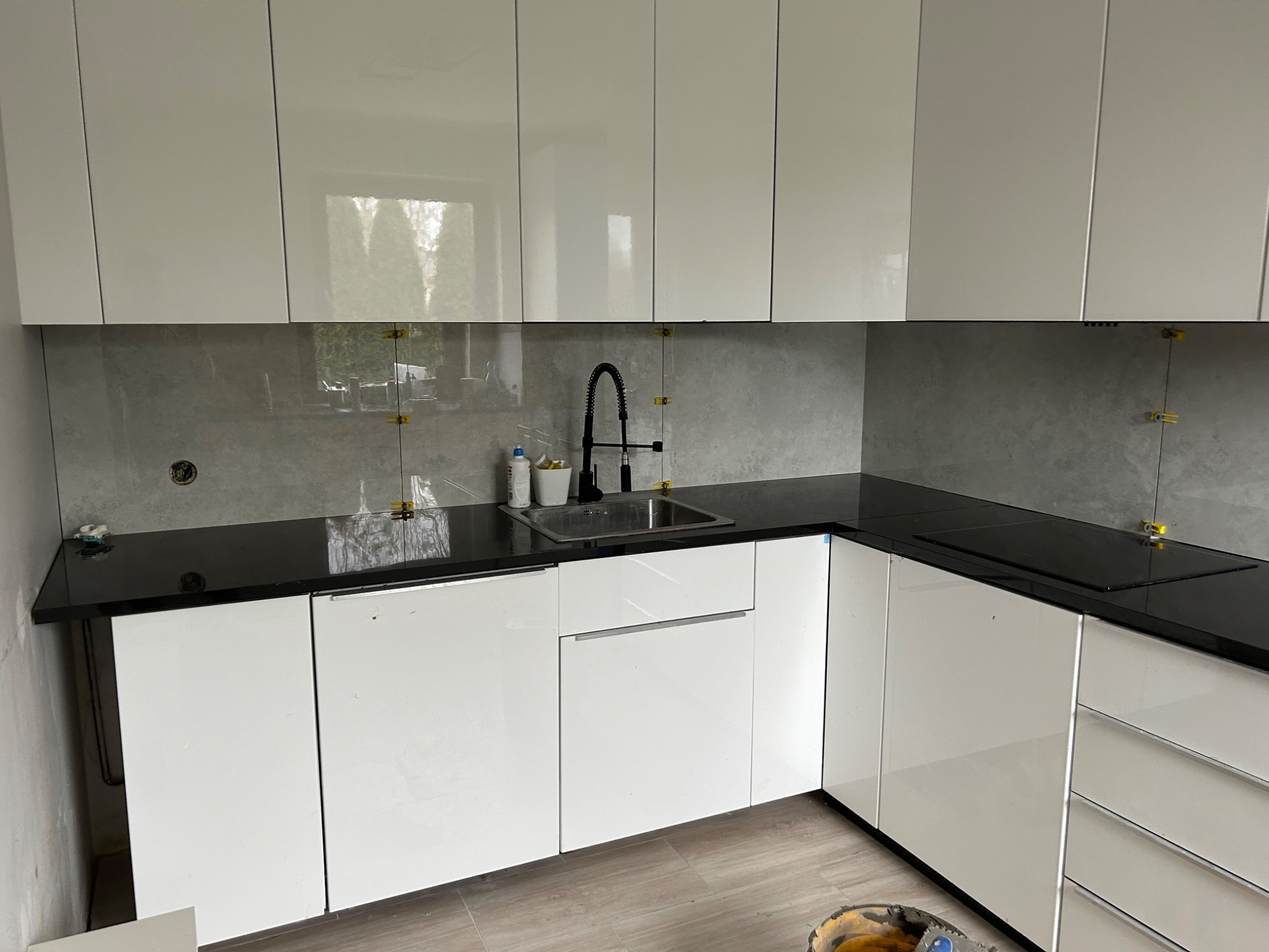 Montaż płytek na ścianie w kuchni w Kamieniu Pomorskim, widoczne białe szafki z czarnym blatem oraz elementy mocujące płytki na szarej ścianie.
