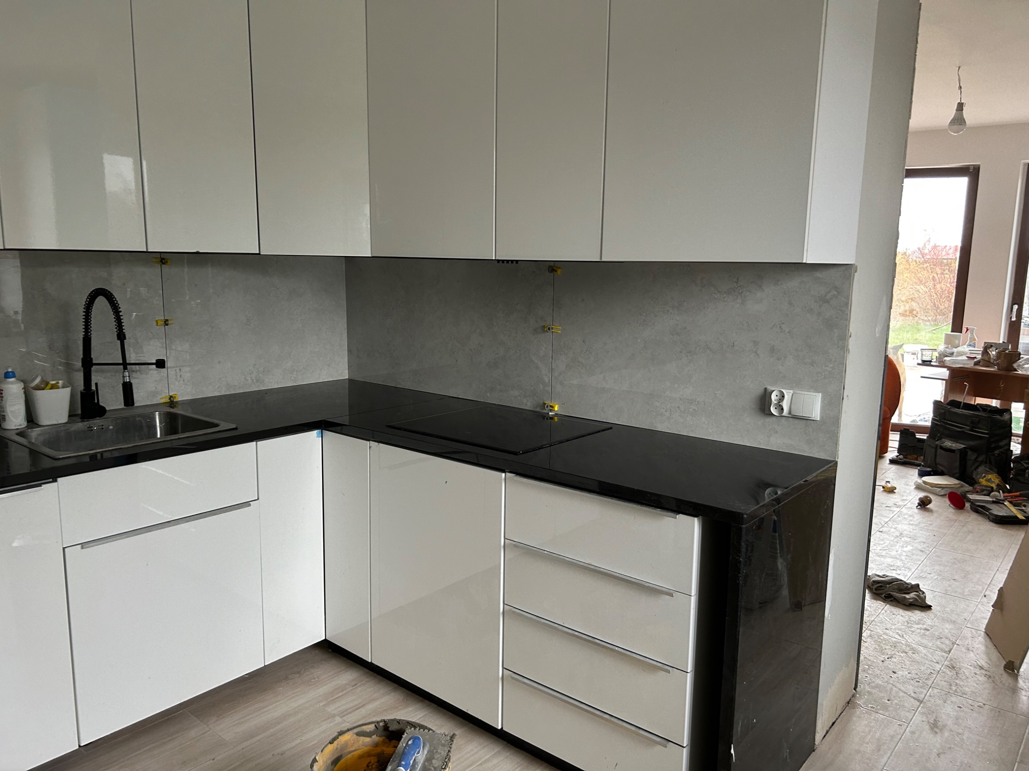 Nowoczesna kuchnia w trakcie wykończenia: białe, lakierowane szafki, czarny blat, szare płytki na ścianie z żółtymi krzyżykami dystansowymi, widoczny sprzęt i narzędzia budowlane na podłodze.