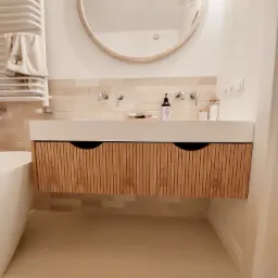 Nowoczesna łazienka z wiszącą szafką pod umywalką z drewnianymi lamelami, okrągłym lustrem z jasną ramą i beżowymi płytkami na ścianie.