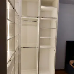 Wnętrze białej, nowoczesnej garderoby z oświetleniem LED, widoczne półki, szuflady i drążki na ubrania, bez zawartości, z elementami metalowymi.