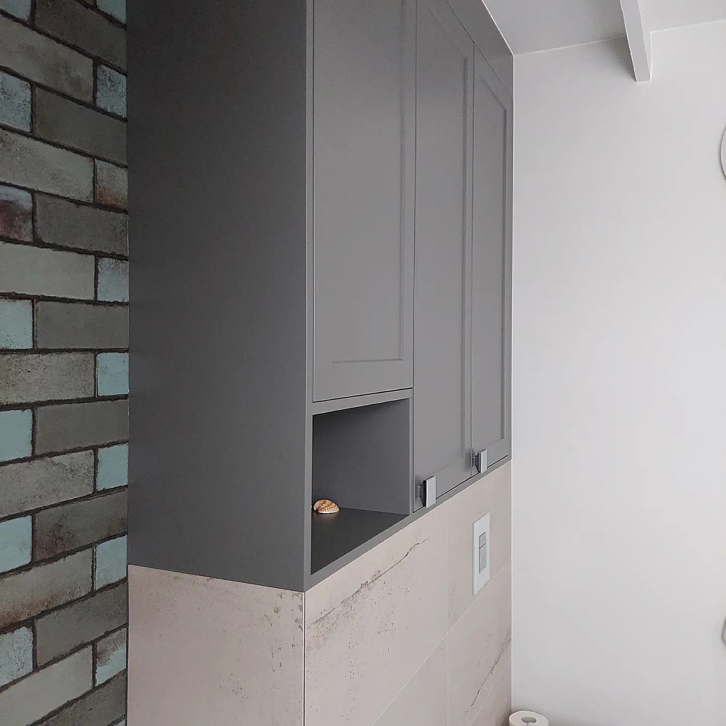 Szara szafka kuchenna wisząca z otwartą półką, na której leży muszelka, w pomieszczeniu wykończonym cegłą i płytkami imitującymi kamień.