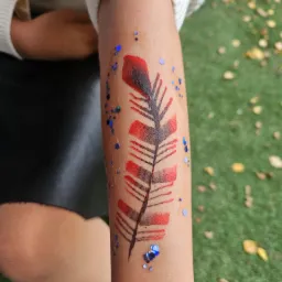 Ręka dziecka z tymczasowym tatuażem przedstawiającym pióro w kolorach czerwieni i czerni, ozdobione brokatem w odcieniach niebieskiego, na tle trawy i opadłych liści.