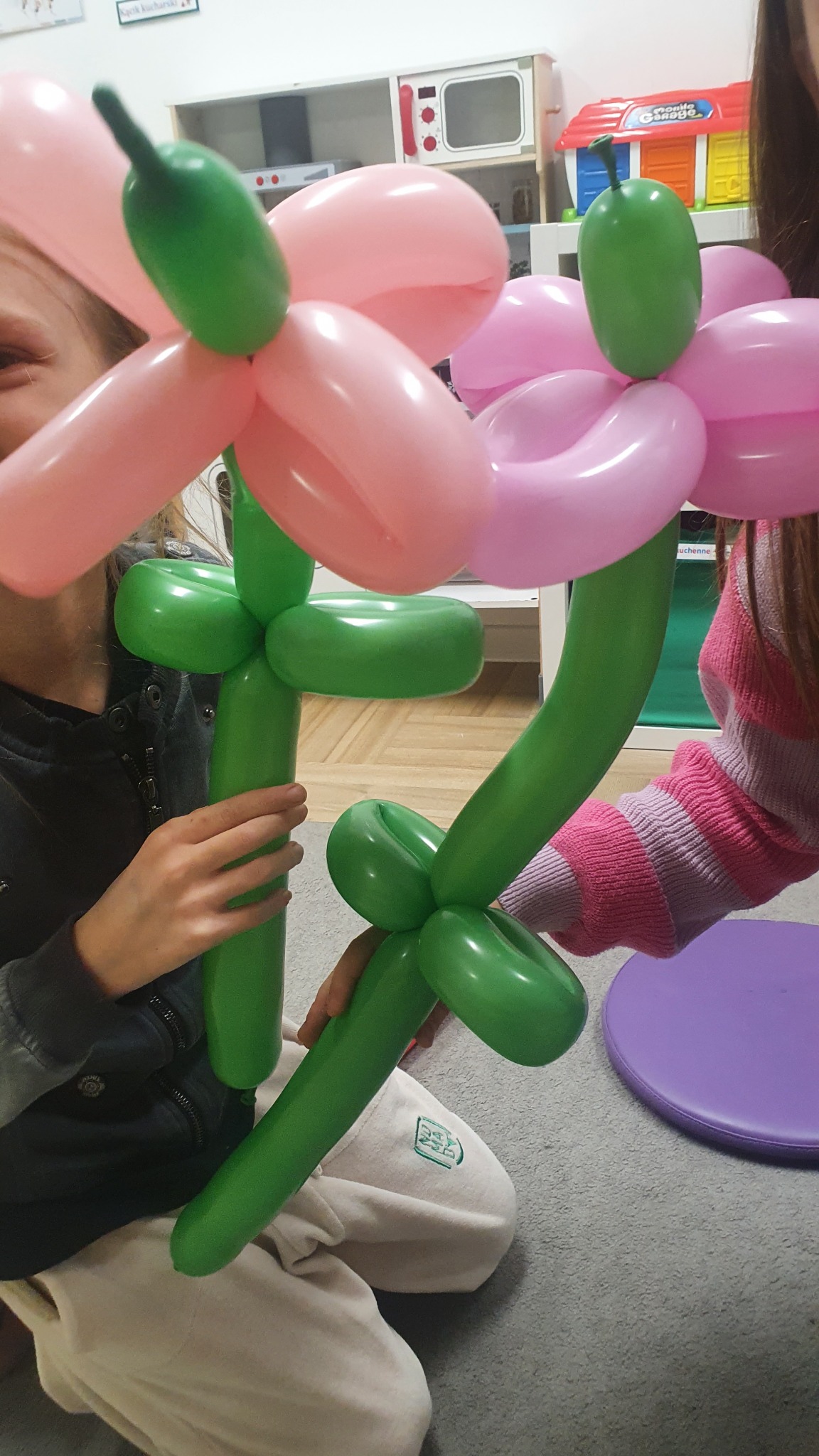Dzieci trzymają w rękach kwiaty wykonane z różowych i zielonych balonów modelowanych, w tle zabawki.