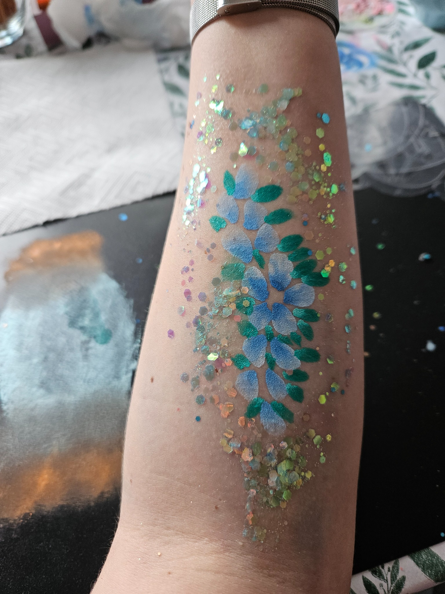 Ramię z tymczasowym tatuażem w kształcie kwiatów, wykonanym z niebieskiej i zielonej farby oraz brokatu w różnych kolorach i kształtach.