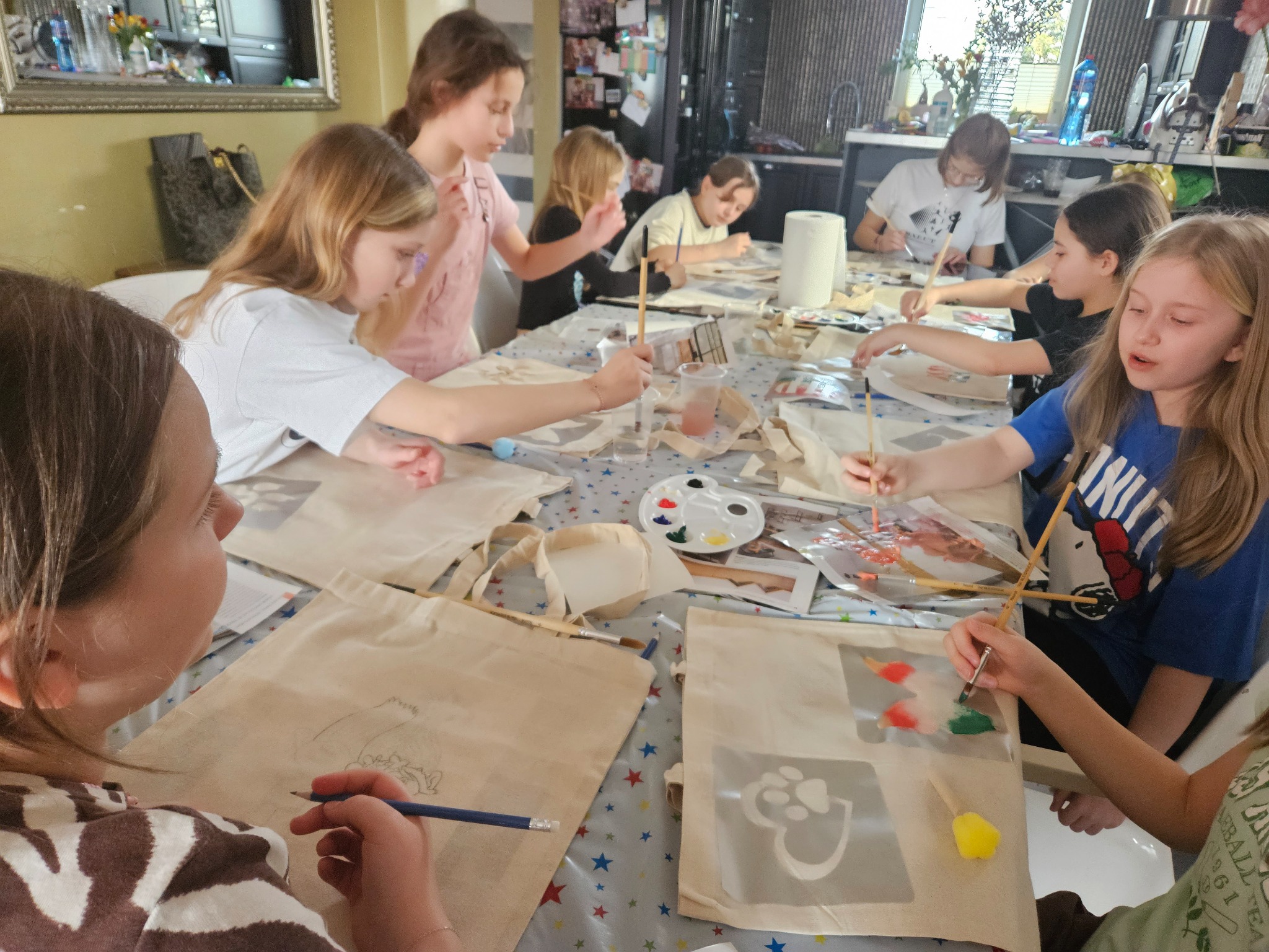 Grupa dzieci maluje farbami na płóciennych torbach pod okiem osoby dorosłej, na stole rozłożone farby, pędzle i szablony