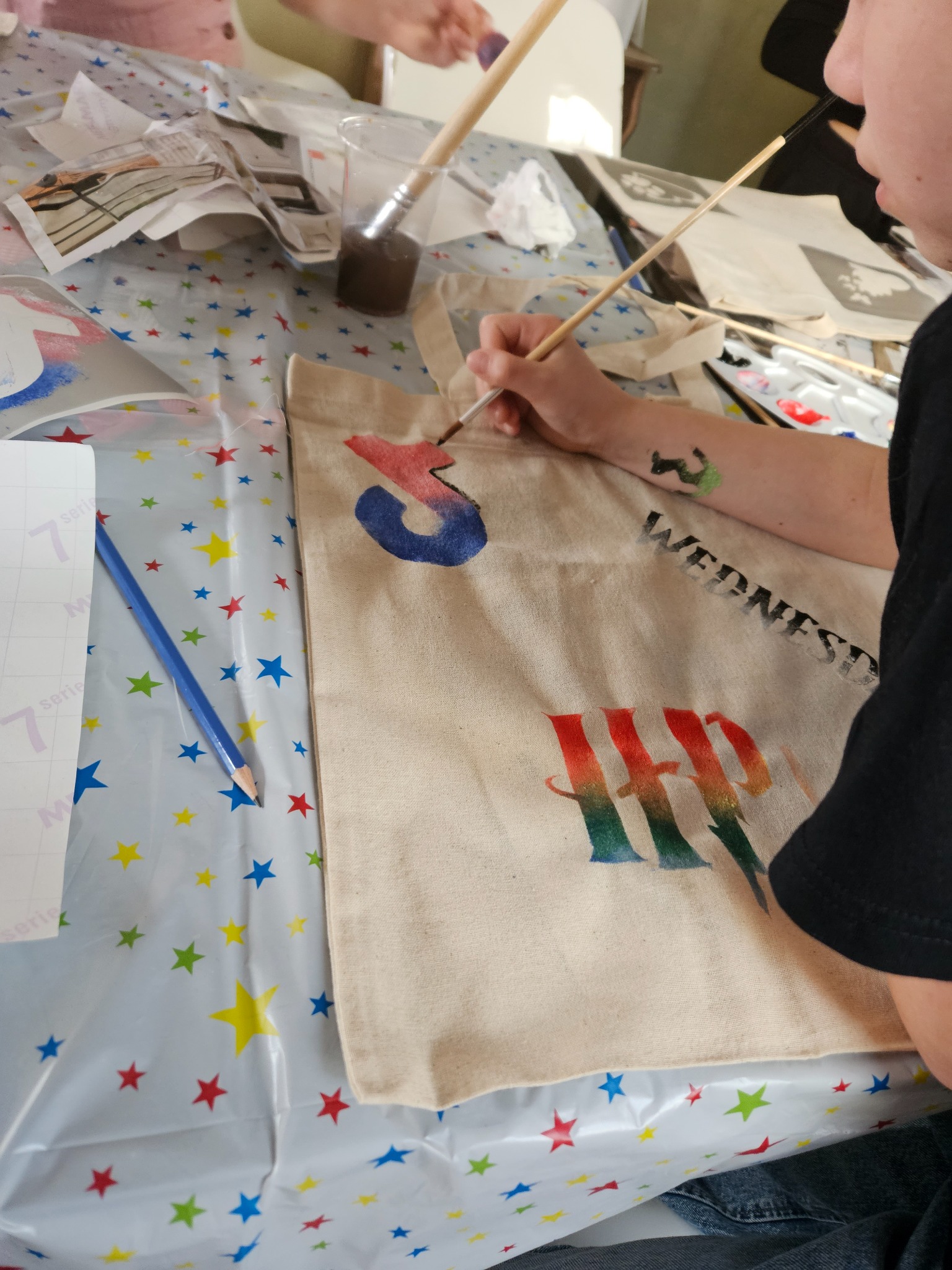 Osoba maluje farbami na beżowej torbie z materiału, na stole z ceratą w kolorowe gwiazdki leży ołówek i pędzle.