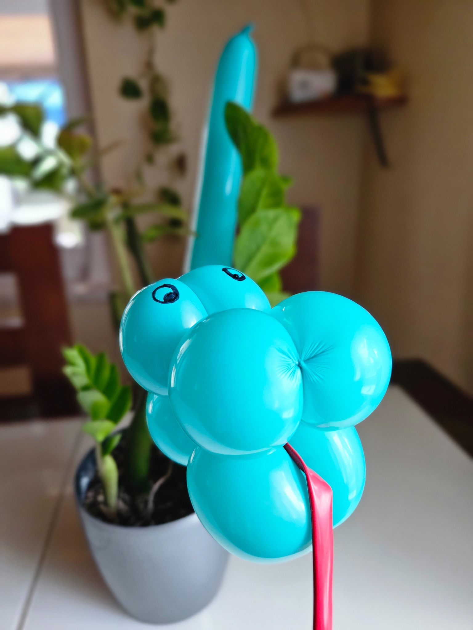 Turkusowy balonowy stworek z namalowanymi oczami, umieszczony obok doniczki z zieloną rośliną na białym stole.