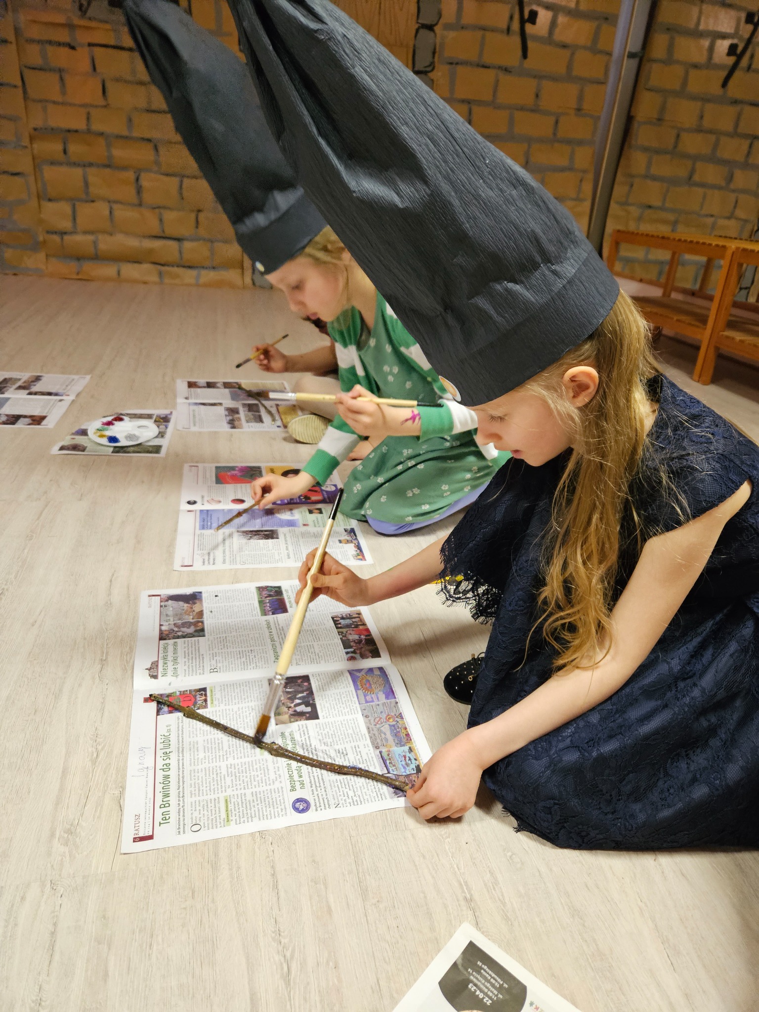 Dwie dziewczynki w wysokich, stożkowych kapeluszach malują farbami na gałązkach leżących na gazetach, siedząc na podłodze w pomieszczeniu o ścianach imitujących cegły.