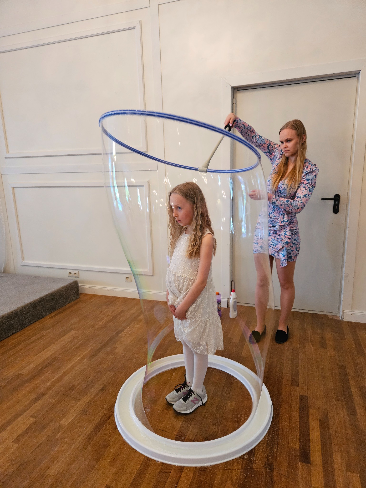 Dziewczynka w białej sukience zamknięta w gigantycznej bańce mydlanej, trzymanej przez kobietę w kwiecistej sukience, na drewnianej podłodze.
