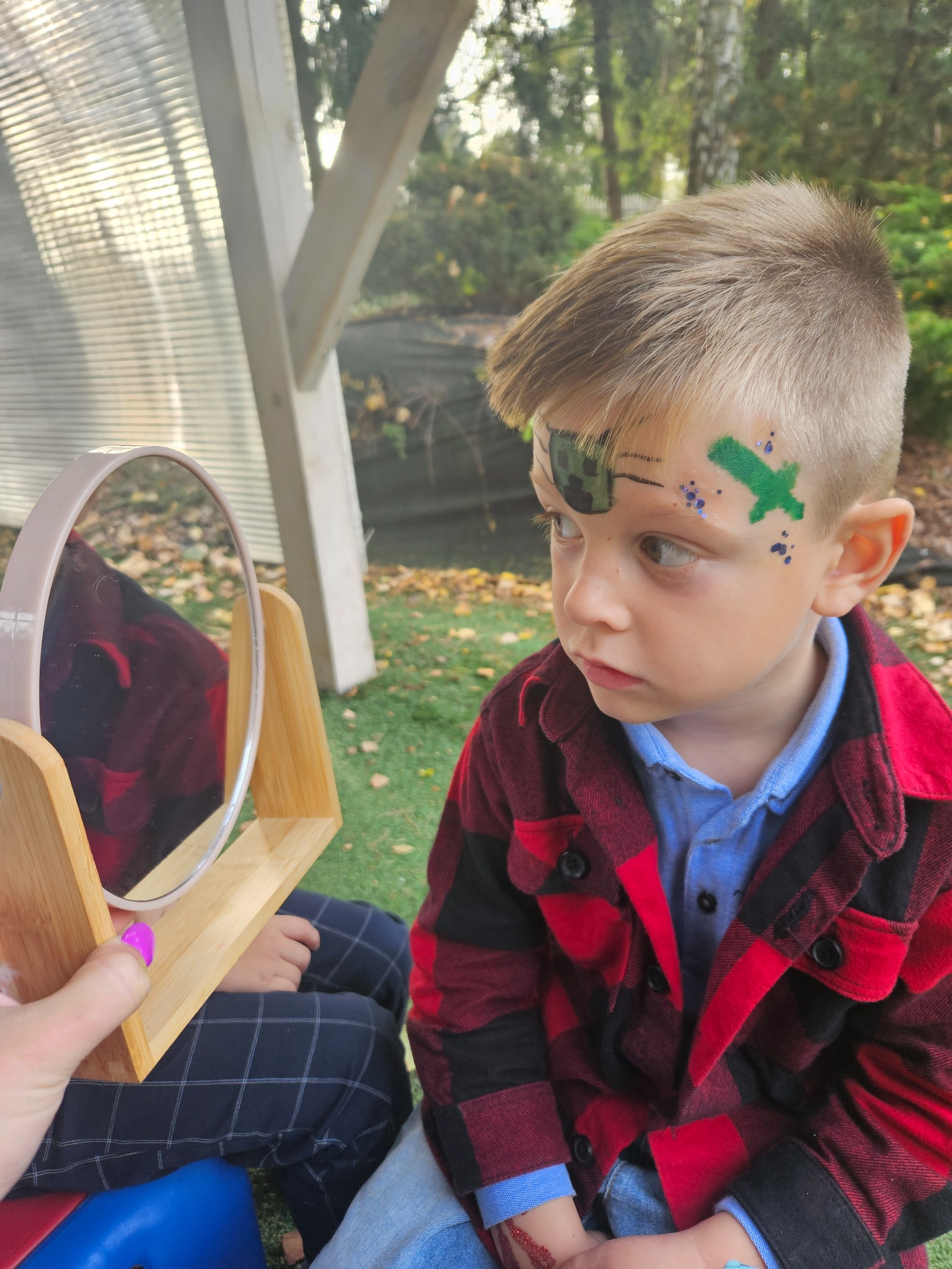 Chłopiec z pomalowaną twarzą w motywy Minecrafta, oglądający się w lusterku trzymanym przez osobę z pomalowanymi paznokciami na różowo, siedzący na tle zielonej trawy i drzew.