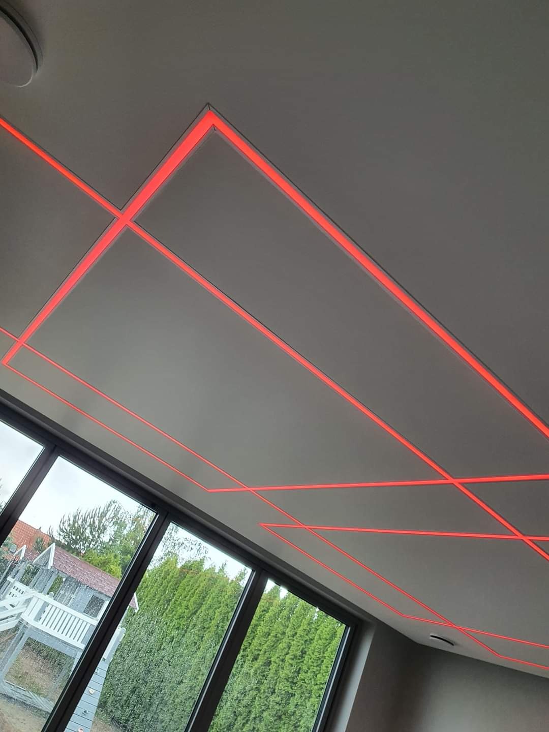 Nowoczesny sufit podwieszany z wbudowanym oświetleniem LED w geometryczne wzory, widok z dołu na tle dużych okien z widokiem na ogród.