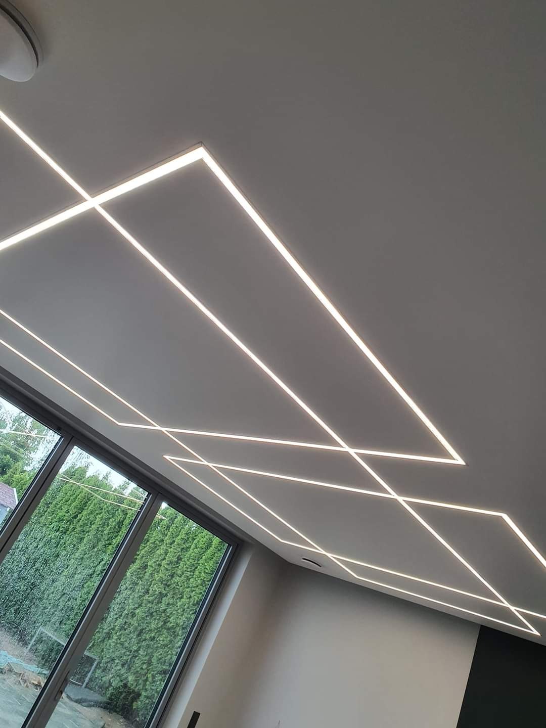 Geometryczny wzór z liniowego oświetlenia LED na suficie podwieszanym w nowoczesnym salonie z dużym oknem.