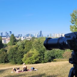 Sprawdzony Detektyw - Aparat fotograficzny na statywie, z widokiem na panoramę miasta i osoby odpoczywające na trawie w parku.