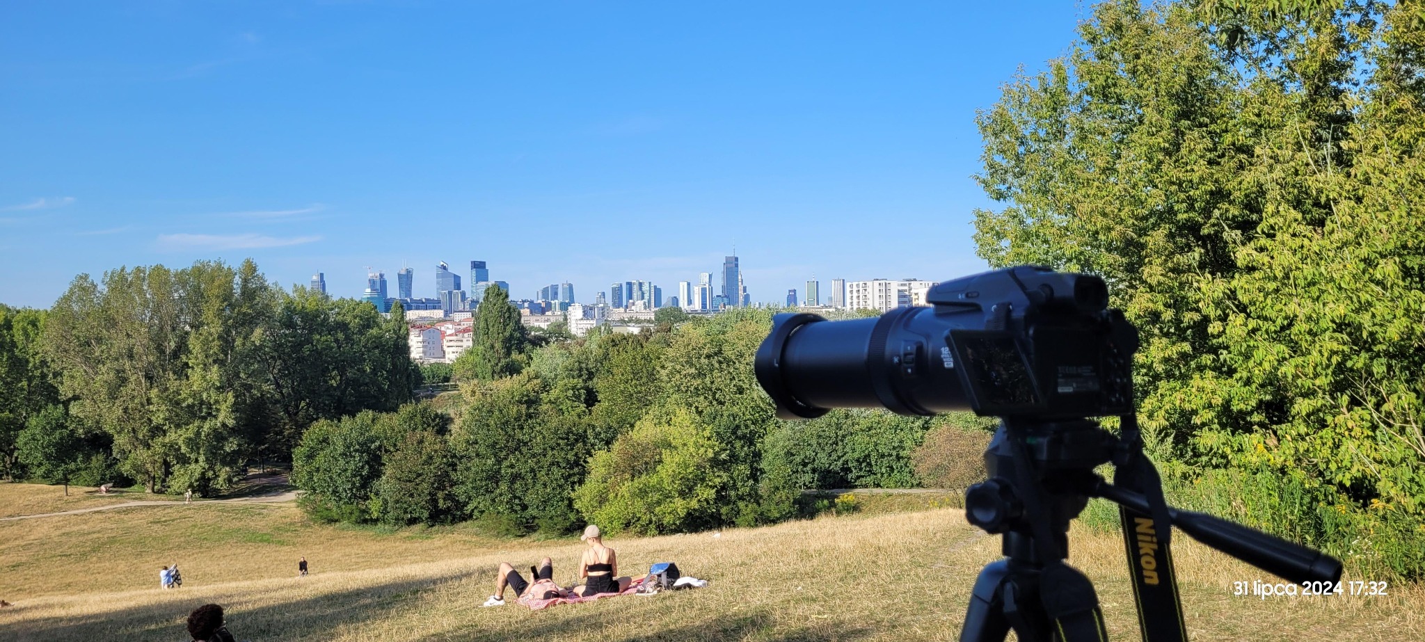 Aparat fotograficzny na statywie, z widokiem na panoramę miasta i osoby odpoczywające na trawie w parku.