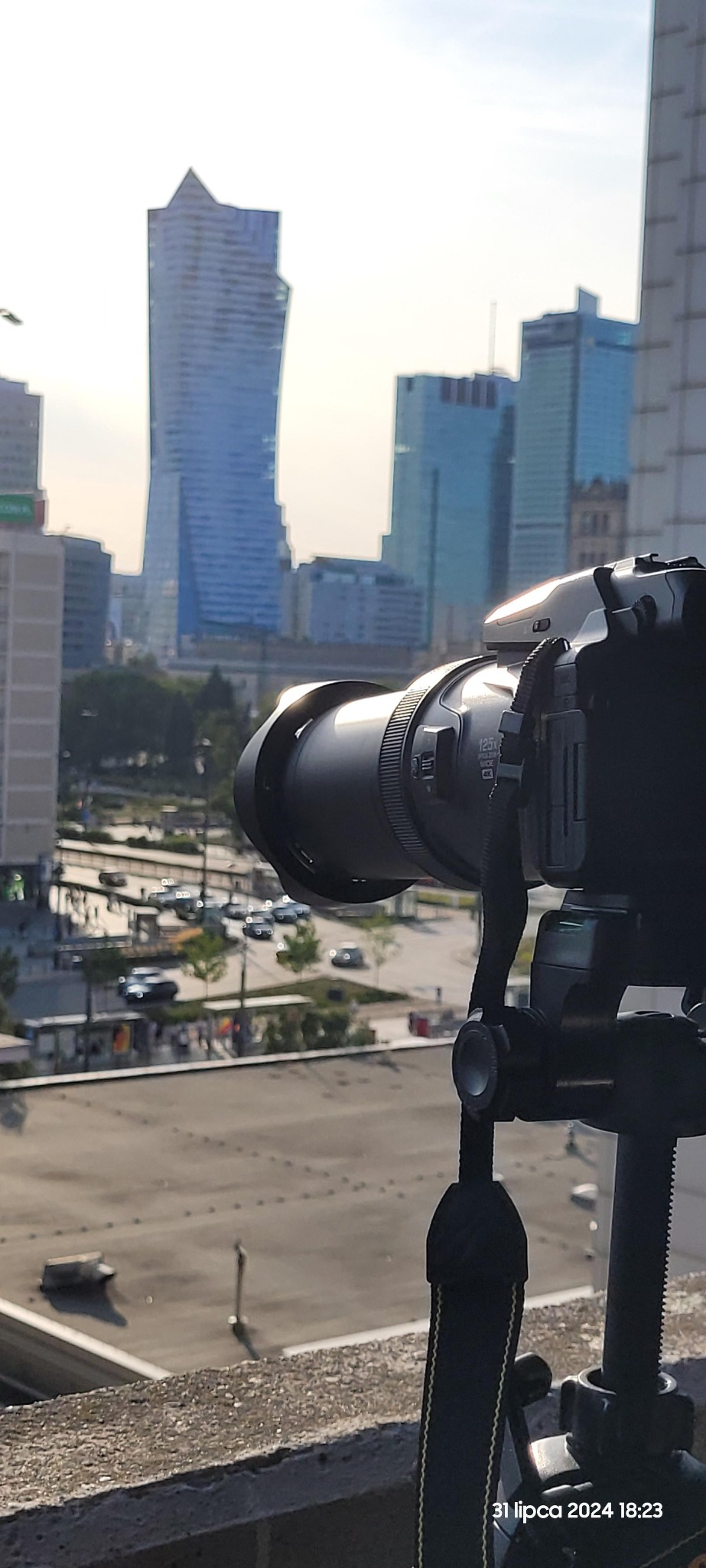 Aparat fotograficzny na statywie, skierowany w stronę panoramy miasta z charakterystycznymi wieżowcami w Warszawie, widoczne ulice i ruch samochodowy.