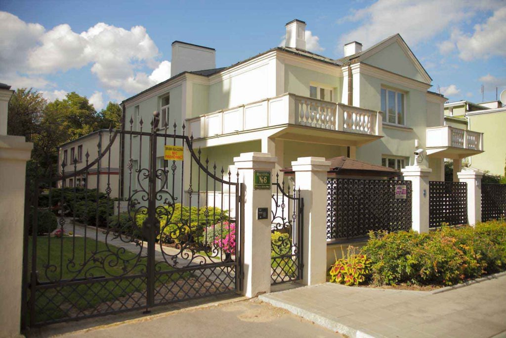 Dwupiętrowa willa w pastelowych kolorach z kutą bramą i ogrodzeniem, tabliczka z numerem posesji przy bramie.