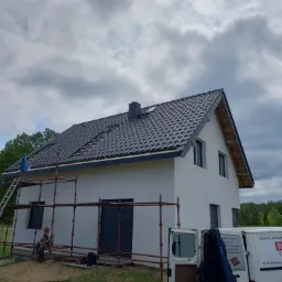 Dom jednorodzinny w trakcie wymiany dachu na czarną dachówkę, widoczne rusztowanie, częściowo zdjęte stare pokrycie, brygada pracowników oraz samochód dostawczy z reklamą firmy dekarskiej.