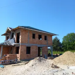 Surowy stan dwupiętrowego domu z czerwonej cegły w trakcie budowy, z widoczną konstrukcją dachu i stertami piasku na pierwszym planie, błękitne niebo w tle.