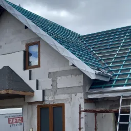 Konstrukcja dachu z łatami i kontrłatami, częściowo pokryta membraną, widoczna z boku domu w trakcie budowy, z oknami i rusztowaniem, przy białym samochodzie firmy dekarskiej.