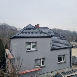Pokrycia Dachowe Paweł Czarnuch - Szary dom jednorodzinny z ciemnoszarą dachówką, widziany z boku, z częściowo widocznym rusztowaniem i samochodem dostawczym firmy dekarskiej. Numer domu 166.