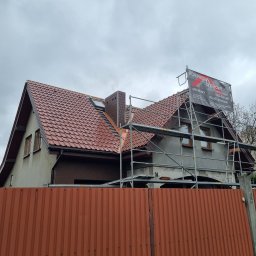 Pokrycia Dachowe Paweł Czarnuch - Dom z nowym, czerwonym dachem z dachówki, widoczny zza pomarańczowego ogrodzenia. Na dachu rusztowanie z banerem firmy dekarskiej.