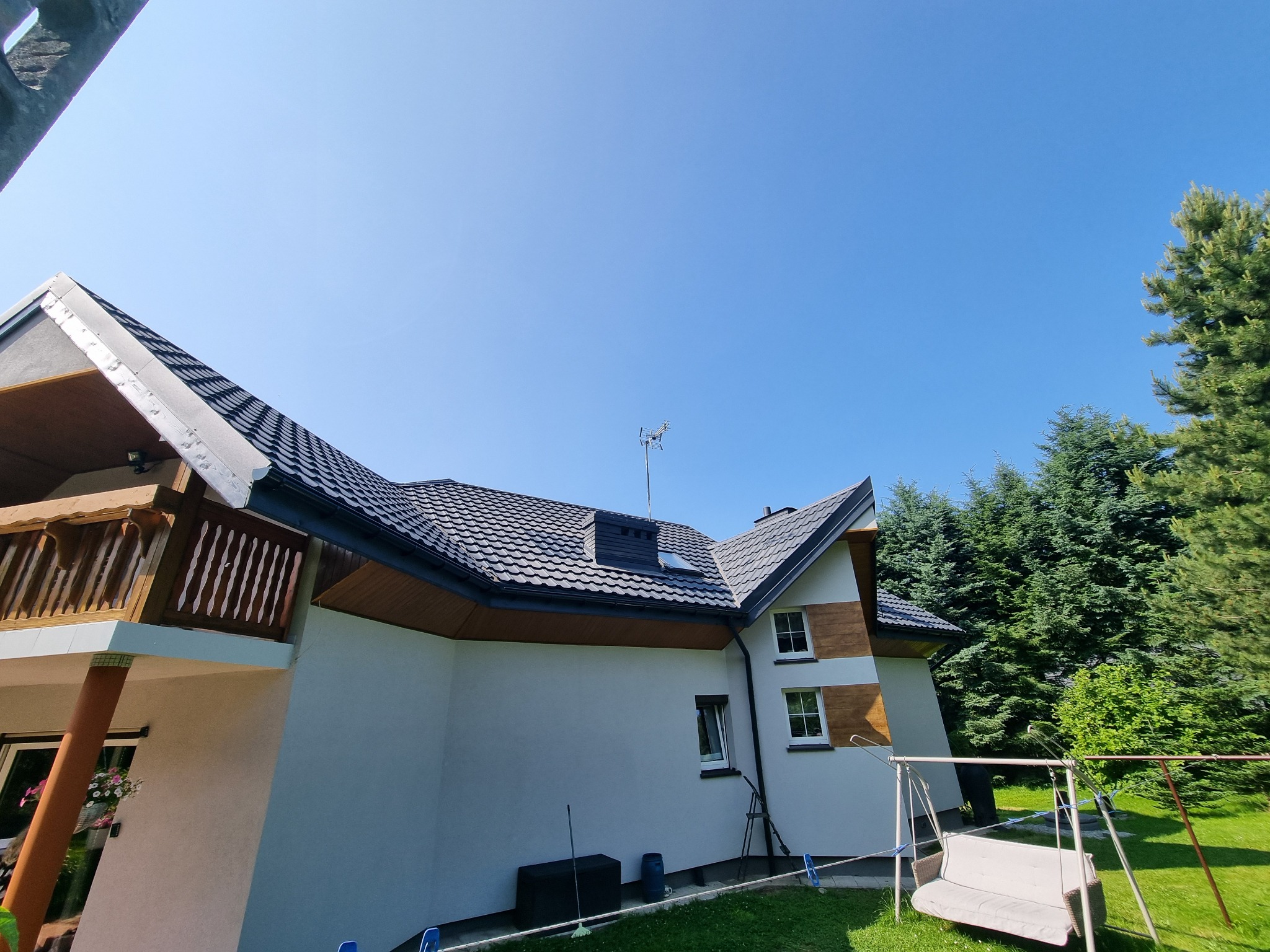Dom jednorodzinny z ciemną dachówką, widoczny z boku, z balkonem i elementami drewnianymi. Na dachu antena i komin. W tle zieleń drzew i błękitne niebo.